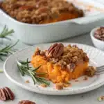 A serving of savory sweet potato casserole with pecans on a plate with rosemary garnish.